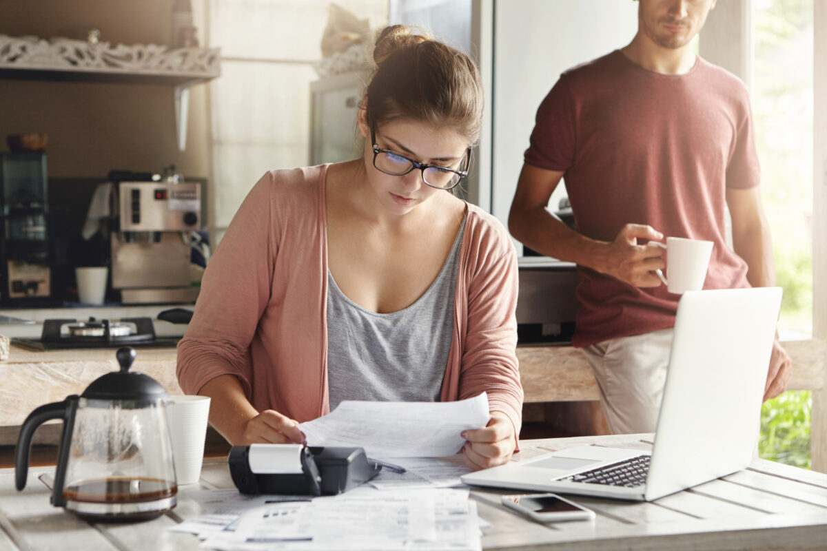 femme assis sur une table avec son ordinateur calculatrice et document pour faire ses comptes. Un homme est debout derrière elle en train de boire un café
