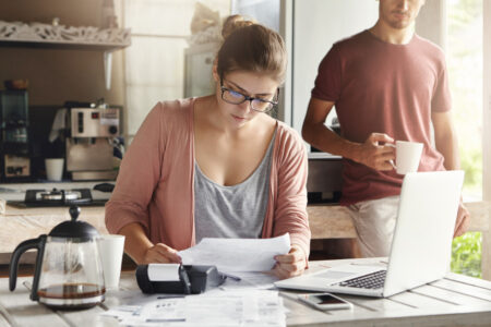 femme assis sur une table avec son ordinateur calculatrice et document pour faire ses comptes. Un homme est debout derrière elle en train de boire un café